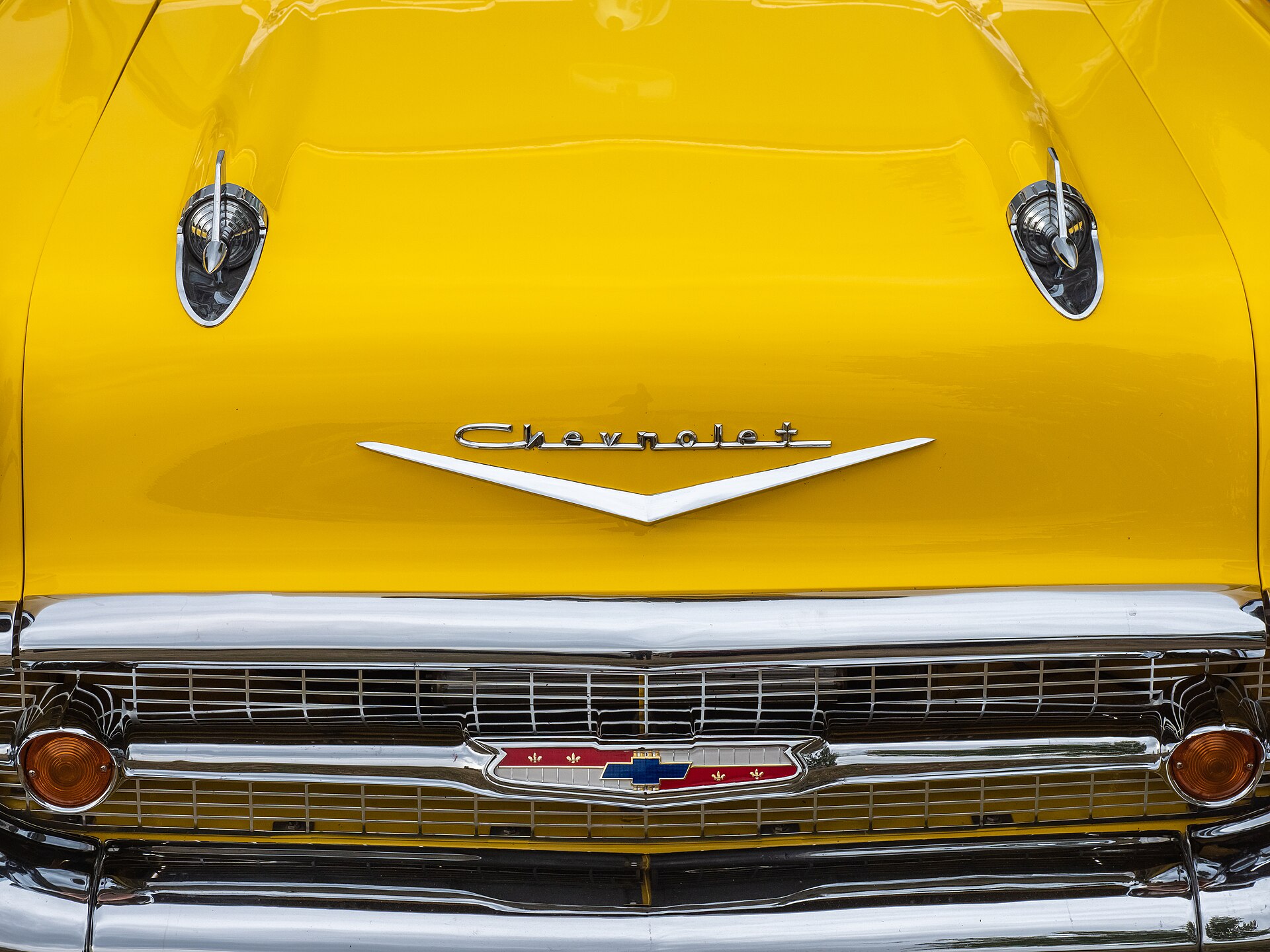 A yellow 1957 Bel Air hood with two hood ornaments, Chevrolet script, a V emblem, and the grille below.